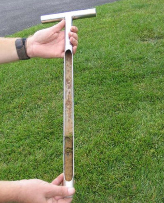 Hands holding a T-shaped soil probe with a visible soil core, used to assess soil conditions prior to sod installation.